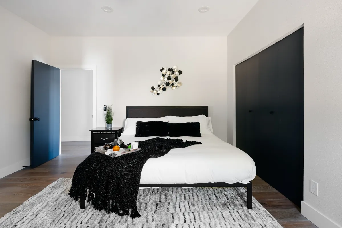 A stylish bedroom with a comfortable bed, elegant wall art, a black wardrobe, and a cozy nightstand. Soft daylight filters in, creating a peaceful atmosphere ideal for relaxation or reading.