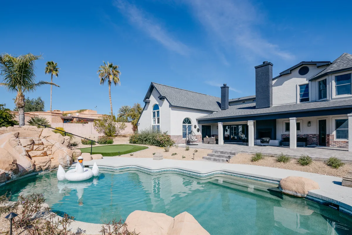 A vibrant outdoor pool scene featuring a sparkling swimming pool, swan float, landscaped rock formations, palm trees, and the house facade in the background. The atmosphere is playful and serene, perfect for group gatherings.