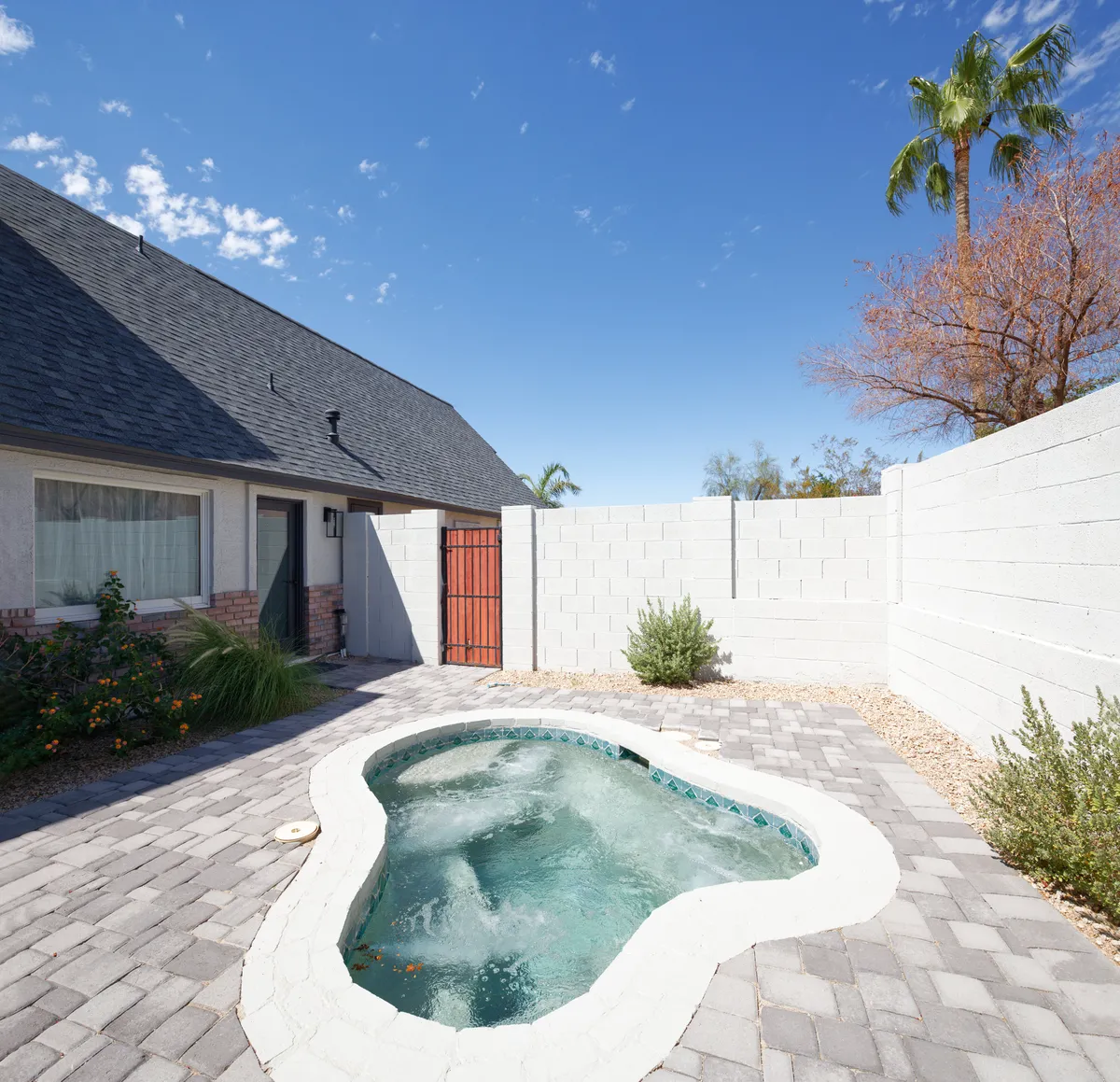 A tranquil exterior spa area with a bubbling hot tub, surrounded by paving, greenery, and a privacy fence. The house is visible in the background, and potted plants enhance the atmosphere of serenity and seclusion.