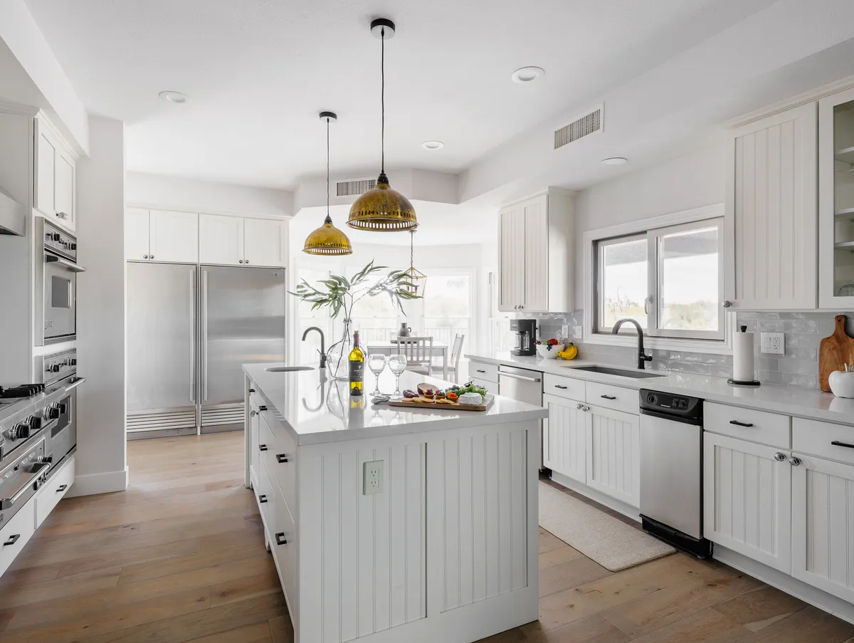 A modern kitchen with an island countertop attractively staged with food, gold pendant lights overhead, large stainless steel appliances, and an open window letting in abundant natural light. The inviting setup suggests both functionality for cooking and a pleasant space for casual dining or entertaining.
