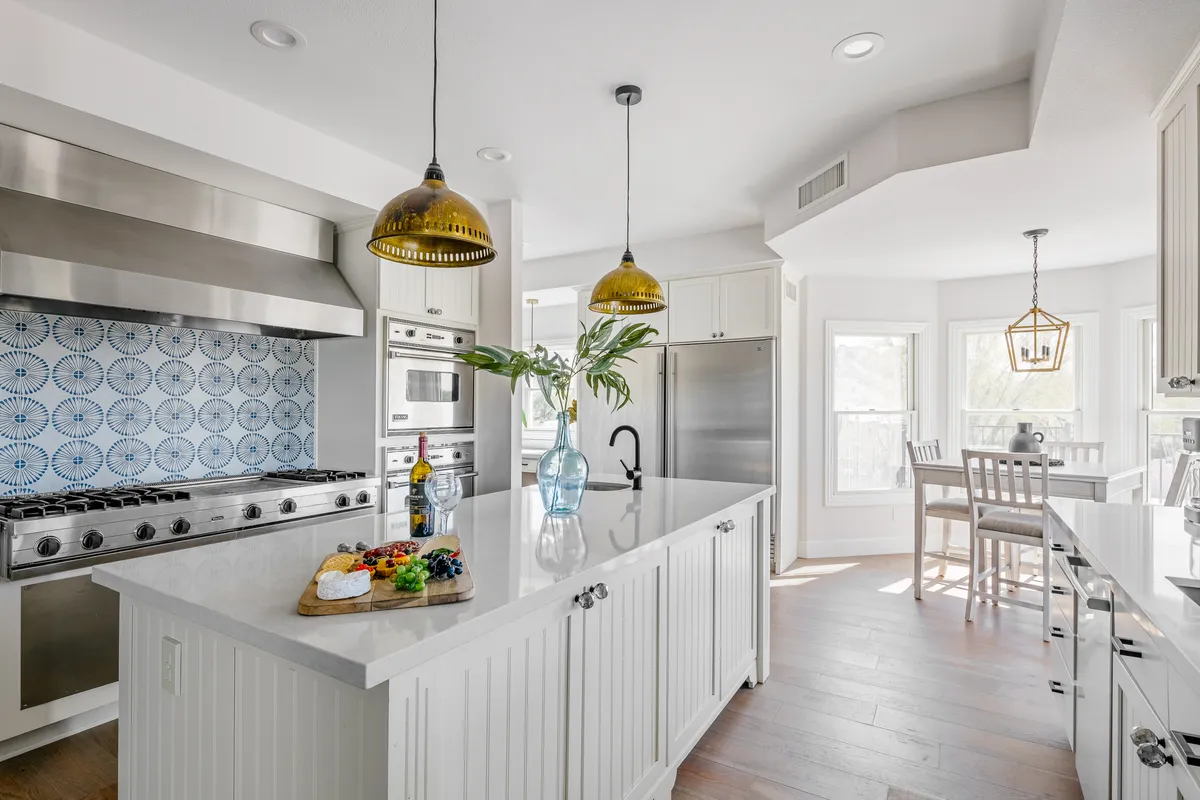 A spacious, cheerful kitchen featuring a central island with bar seating, stainless steel high-end appliances, a striking blue backsplash, and modern pendant lighting. The kitchen is bathed in natural light, and there's visible space for meal prep and gathering, suggesting both culinary functionality and a welcoming, social atmosphere.