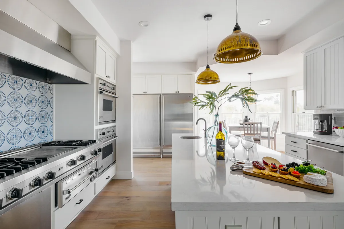 A lively kitchen scene with a large gas stove, a central island topped with a charcuterie board, stylish pendant lighting, and expansive windows. The space is designed for both cooking and socializing, with clear zones for meal prep and conversation.