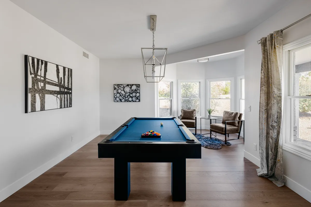 A stylish game room with a central pool table, modern artwork on the wall, and armchairs set by a sunlit window, inviting social interaction and friendly competition.