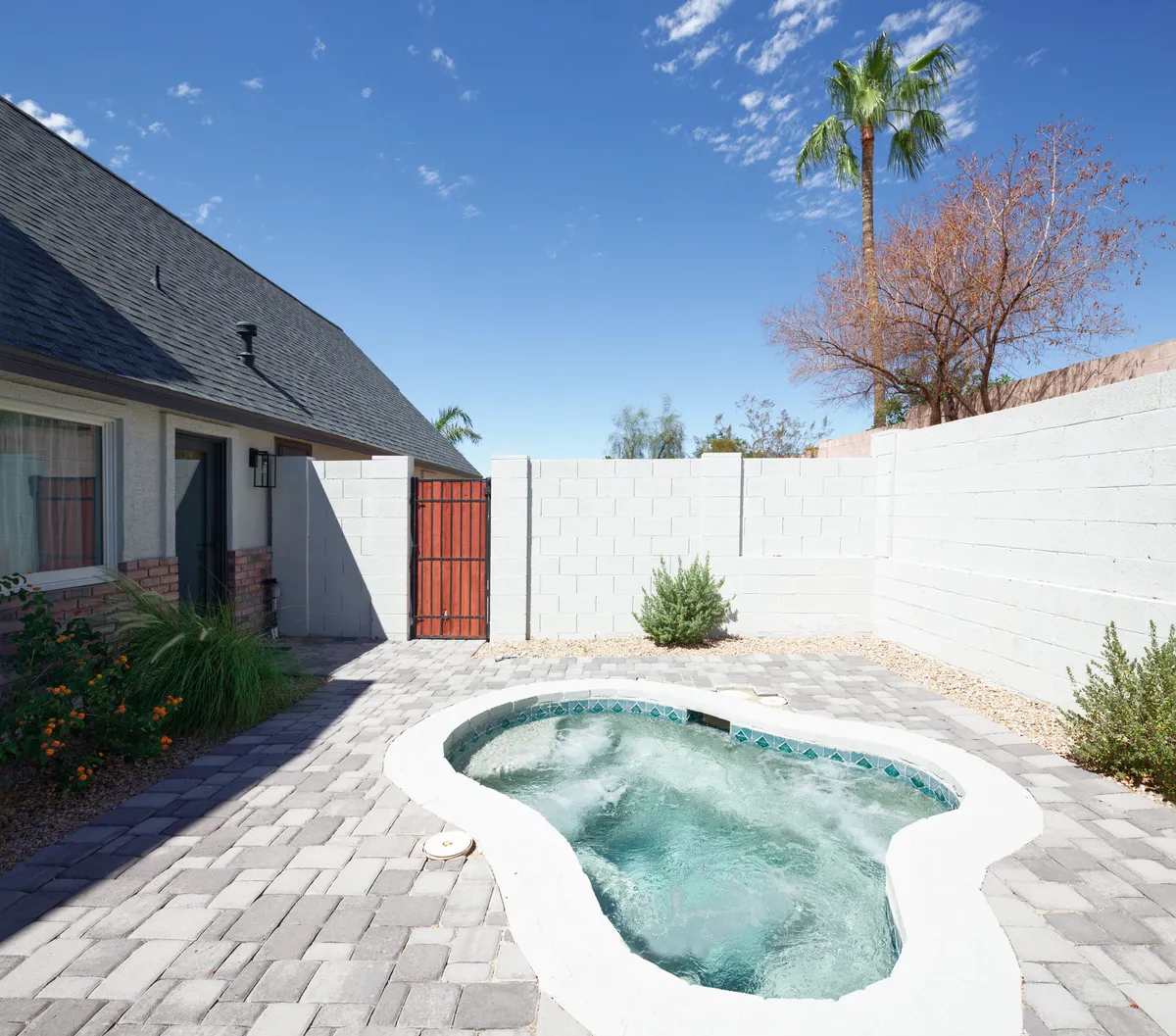 An elegant exterior scene featuring an outdoor dining area set beside a landscaped garden and a hot tub, all against the brick facade of the house. The patio is arranged for relaxing meals and social gatherings, with greenery providing a tranquil backdrop.