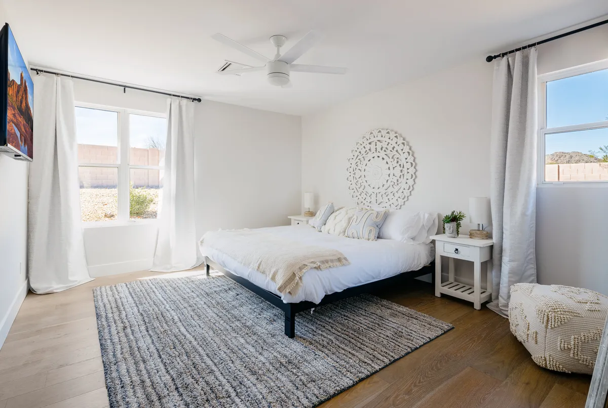 A serene bedroom scene with a textured bed, decorative wall art, a large window letting in abundant natural light, and a TV mounted on the wall. The bedding looks plush and inviting, while the overall ambiance is cozy and quiet.
