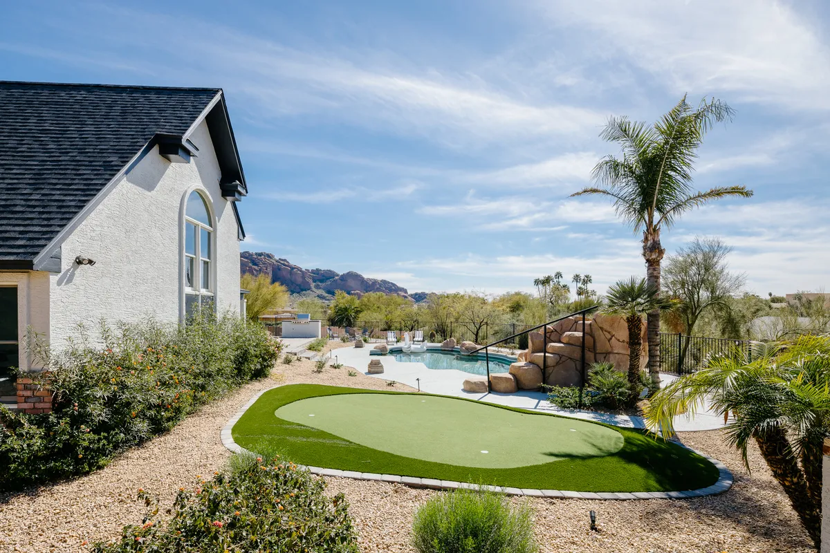 A vibrant exterior scene showcasing a sparkling swimming pool, a well-manicured putting green, lush landscaped gardens, and a stunning mountain view in the background. Palm trees line the scene, and the area is set up for both relaxation and outdoor activities, suggesting a luxurious and playful atmosphere.