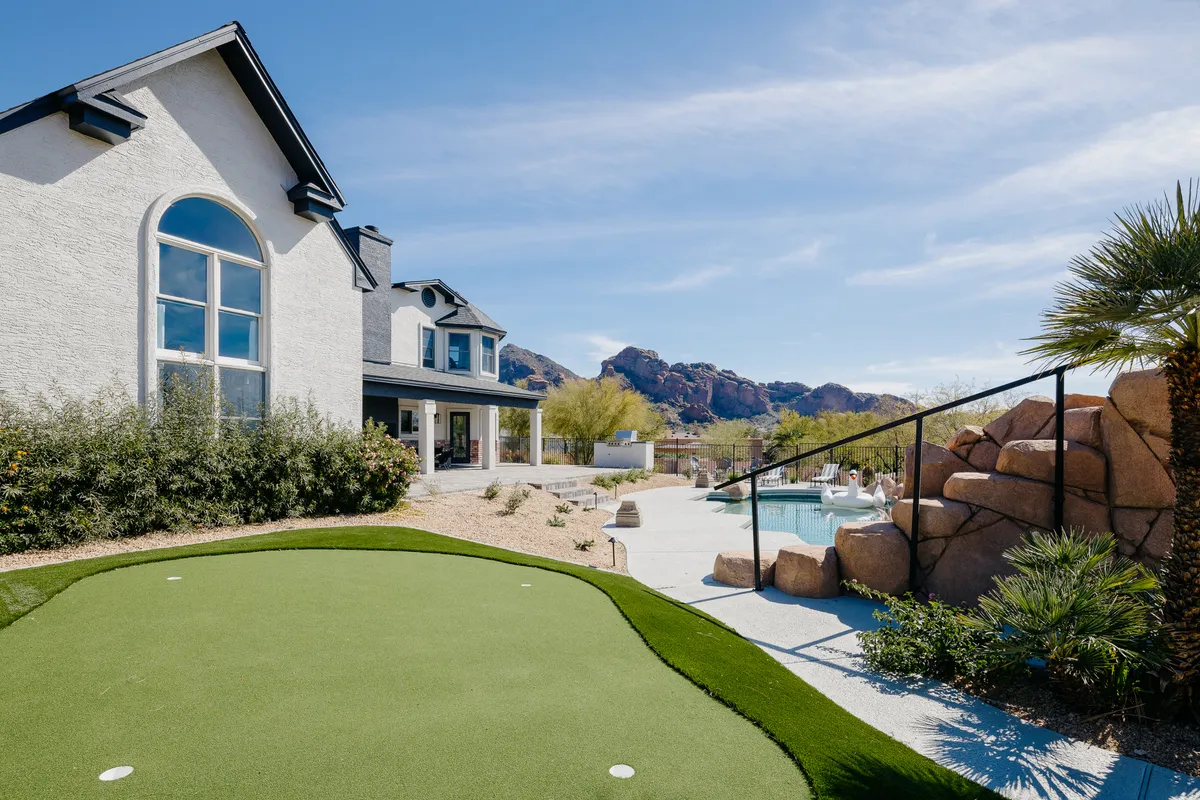 A beautifully landscaped backyard with a putting green, swimming pool, mountain views, and lush gardens. The layout offers space for golf practice, relaxation, and outdoor gatherings, all under a colorful sky.